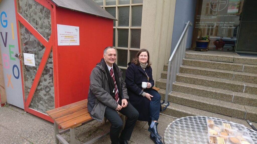 Auf der neuen Sitzbank vor der Friedenskirche sitzen Pastor Holger Panteleit und Laiendelegierte Isolde Dittrich. Dahinter sieht man die Givebox.